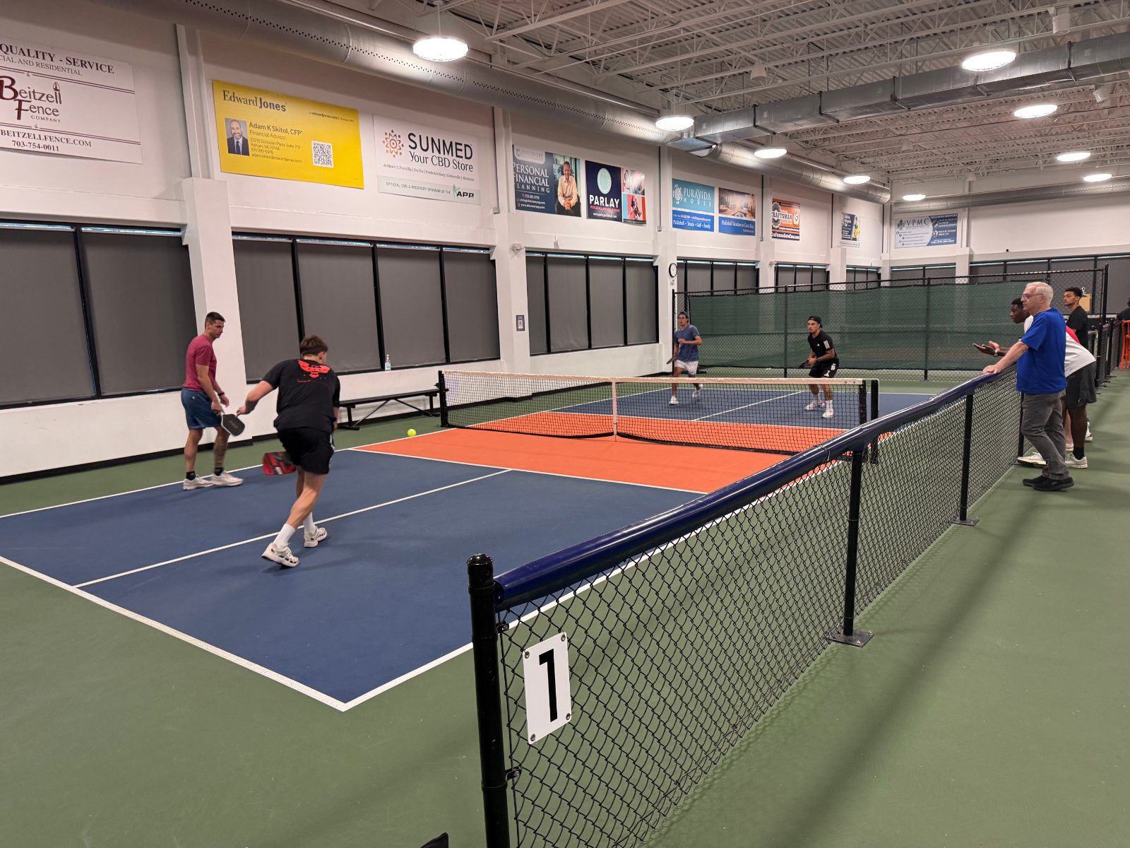 Indoor play on court 1 — permanent nets, full-height divider fencing between courts, even overhead lighting.